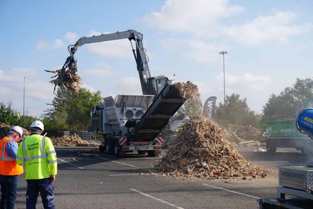 A shredder being used to shred wood waste into a pile. Two men in hi-vis can be seen infront.