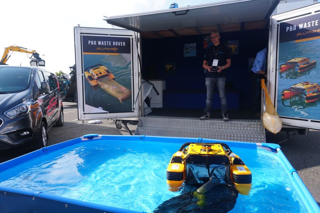 A demonstration of P&D's JellyfishBot being used to collect tennis balls in a small pool in front of a promotional stand. Inside the stand, a man is used a remote control to control the device.