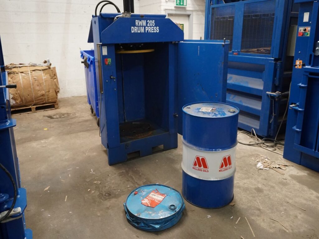 two steel drums, one has been compacted, in front of a drum press machine