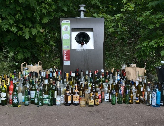 A glass recycling bin with bottles stood in front of it. 